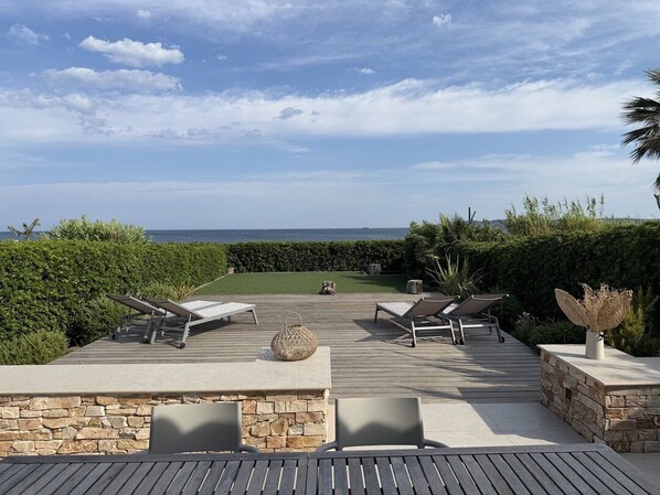 A deck with lounge chairs and a table, overlooking a landscaped garden and the sea.
