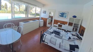 A compact room with a bed, a small table, a chair, a radiator, and a window with a view of trees and buildings. - Studio for 2 people in the heart of Saint-Tropez (ST TROPEZ)