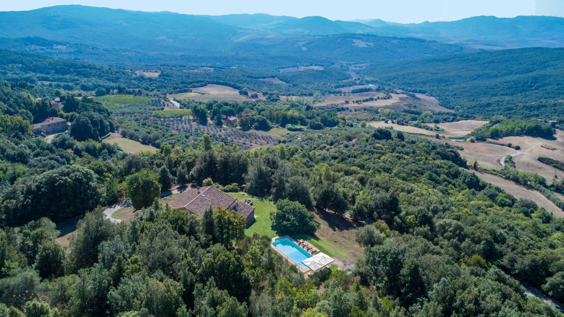 An aerial view of a property with a swimming pool surrounded by dense forest, a large building, and rolling hills in the background.