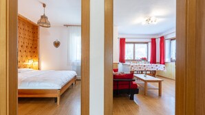 A room with a bed, a wooden headboard, a bedside lamp, a window with curtains, and a view of a dining area with a table and chairs. - Majon Fior de Bosch 1 (Campitello di Fassa)
