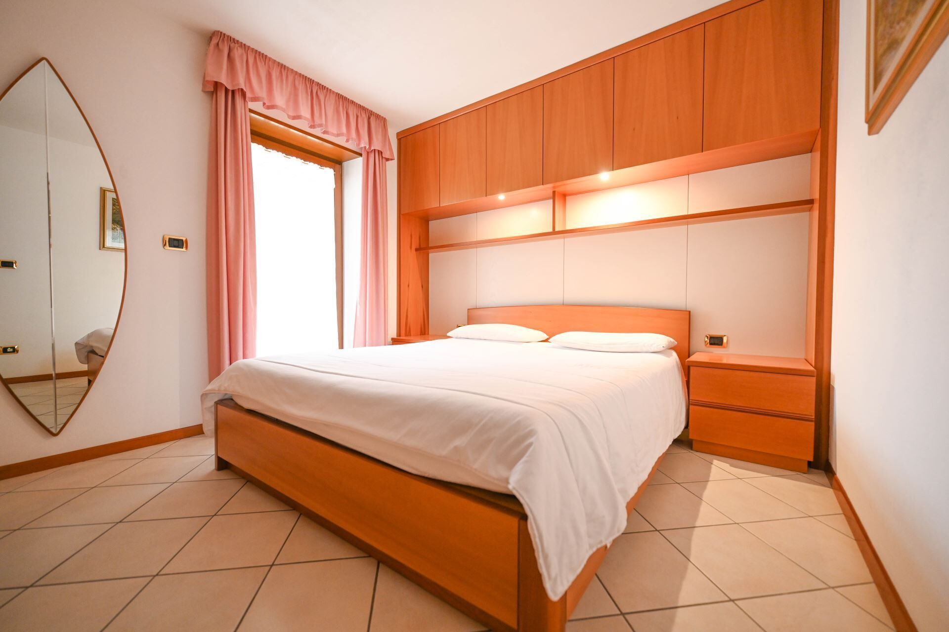 A bedroom with a wooden bed, a nightstand, a mirror, and a window with pink curtains.