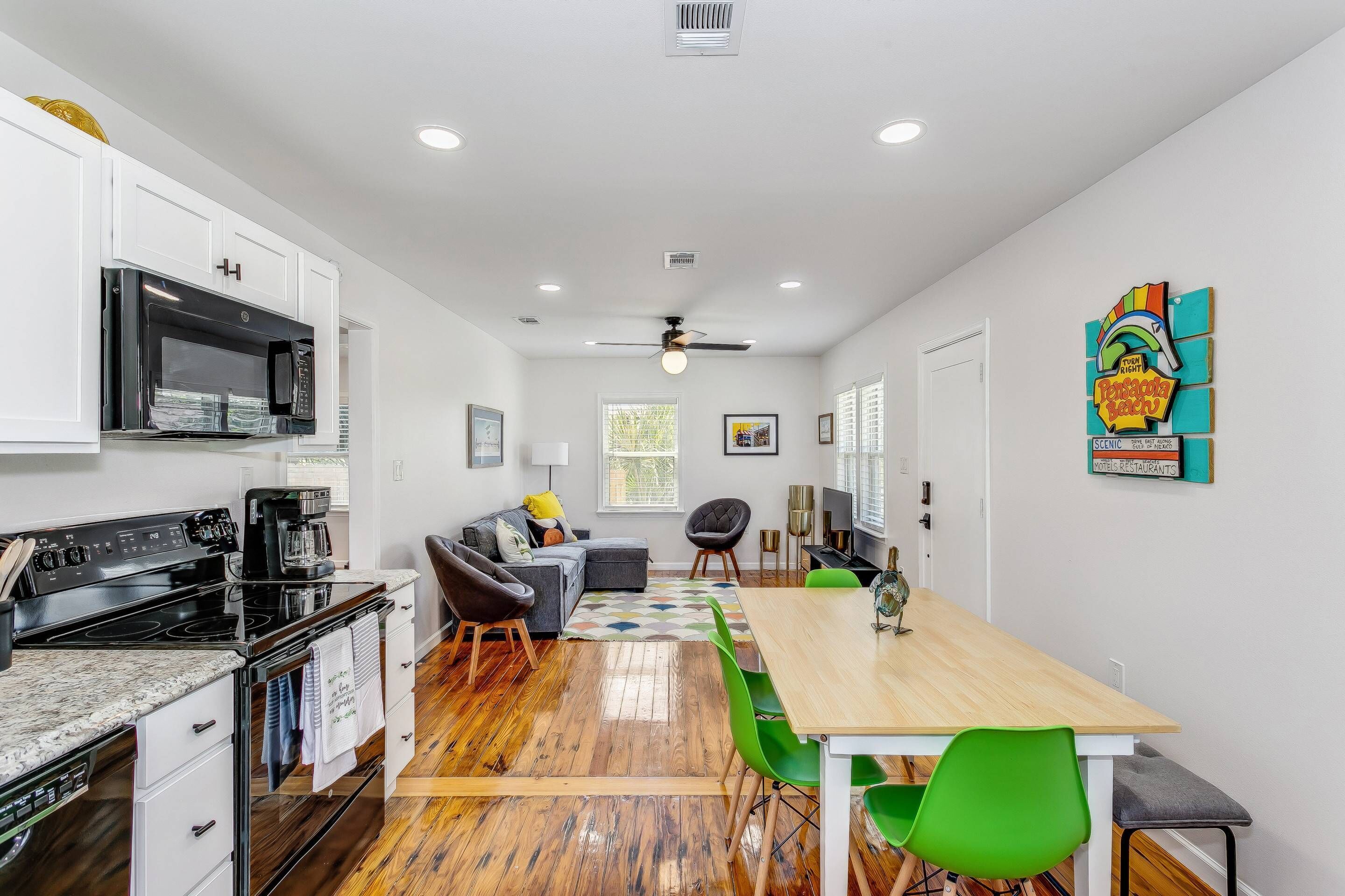 A modern kitchen with white cabinets, a black microwave, and a stove. There is a dining area with a wooden table and green chairs.
