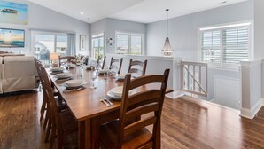 A dining room with a wooden table set for six, surrounded by chairs. There are windows with blinds, a chandelier, and a view of the outside. - Amazing Home with Pool, Golf Cart, Elevator, Hot Tub, Fire Pit Short Walk to the Beach (Oak Island)