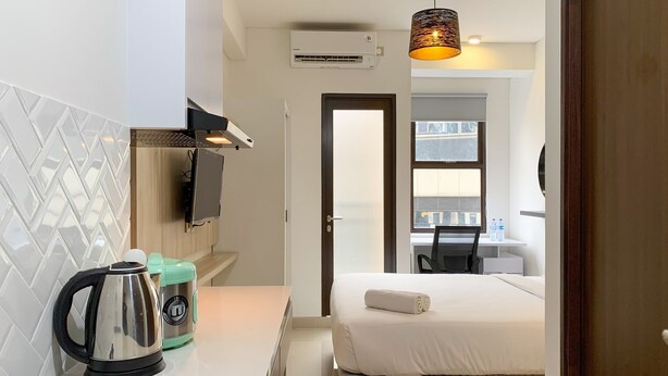 A compact hotel room with a kitchenette, a bed with a rolled towel, a hanging lamp, and a view through a door. - Elegant Studio At Transpark Cibubur Apartment Near Shopping Mall (Bekasi)