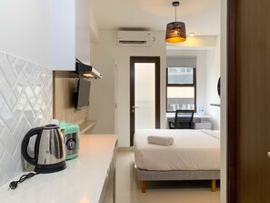 A compact hotel room with a kitchenette, a bed with a rolled towel, a hanging lamp, and a view through a door.