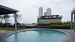 A swimming pool with a curved edge, surrounded by a paved deck and greenery. In the background, there are high-rise buildings and a body of water.