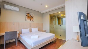 A modern hotel room with a bed, a desk, a chair, a TV, and a glass door leading to a bathroom.