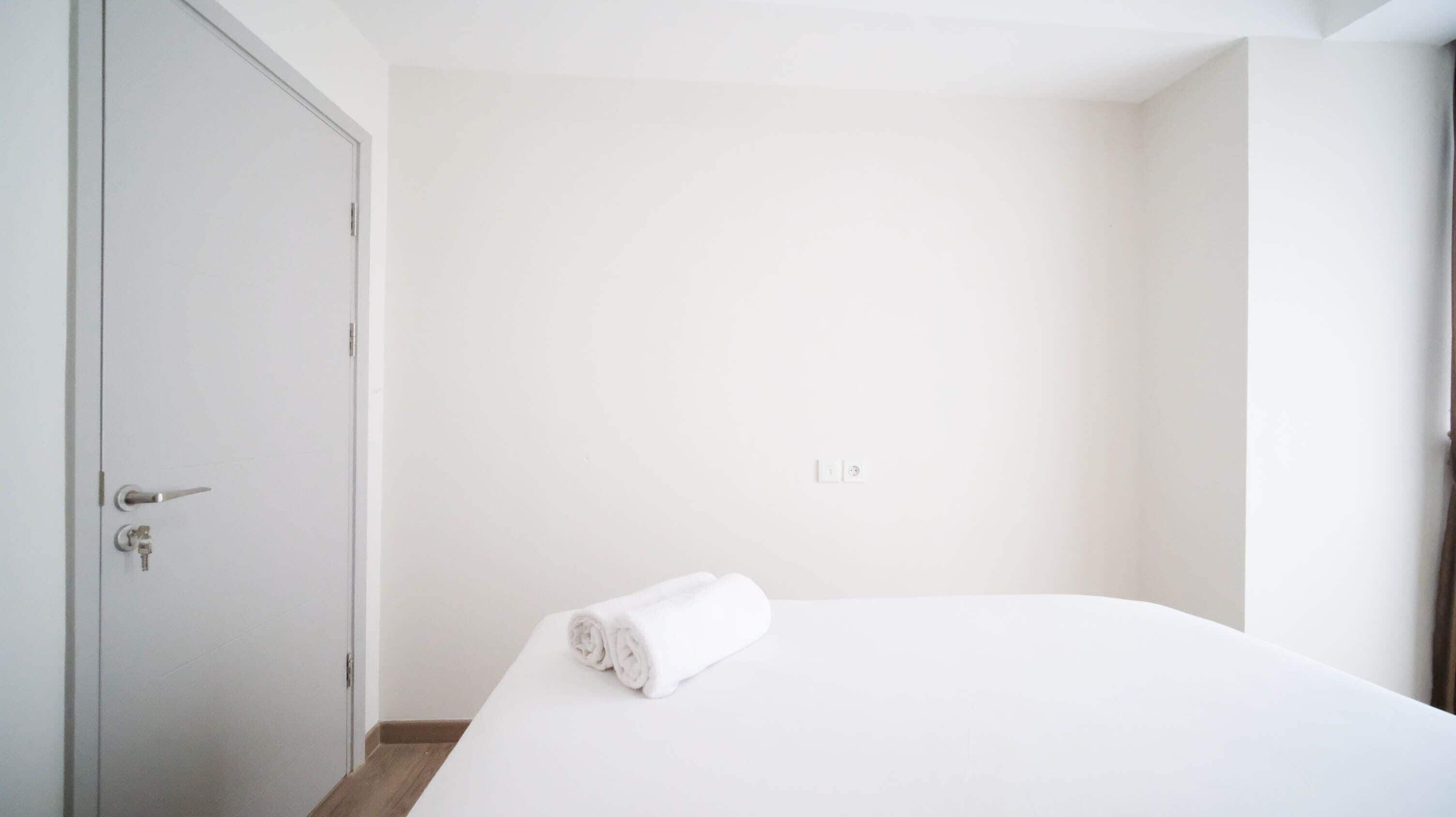 Minimalistinen hotellihuone, jossa on yksi sänky, ovi ja pieni pyyhe sängyllä.