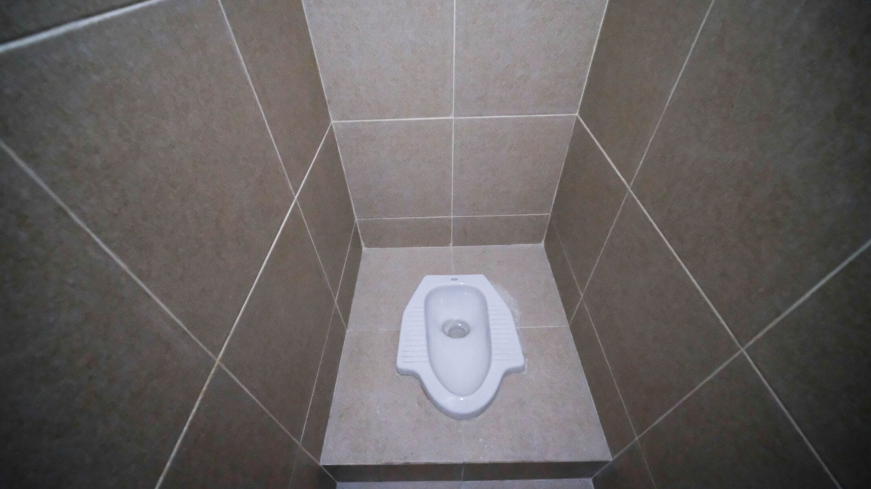 A small, square-shaped toilet with a white seat and a central drain hole, surrounded by brown tiles.