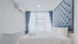 A modern hotel room with a large bed, a tufted headboard, a window with blue curtains, and an air conditioning unit on the wall. - Comfort Designed 1Br At Brooklyn Alam Sutera Apartment (South Tangerang)