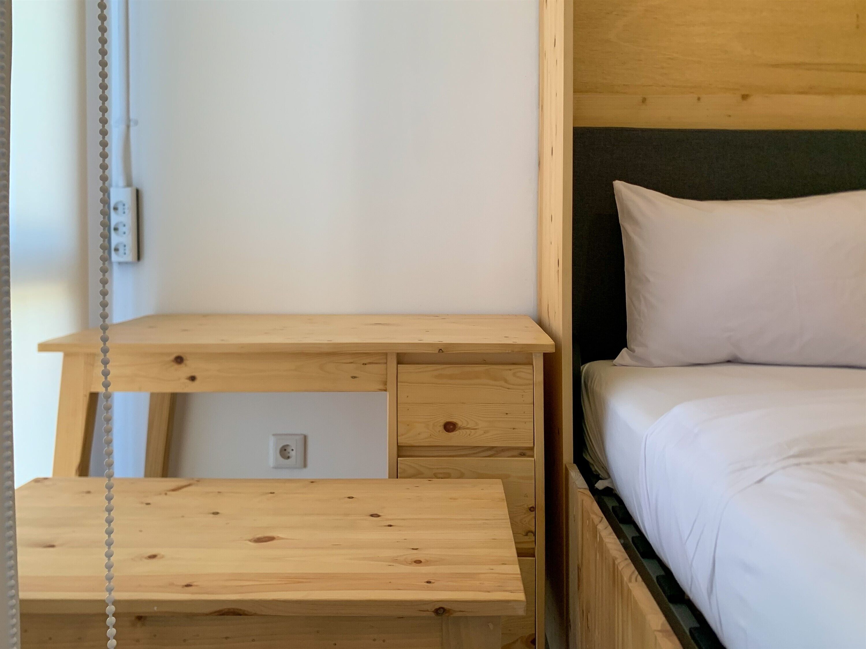 A wooden bed with white bedding, a wooden nightstand with a drawer, and a wall socket.