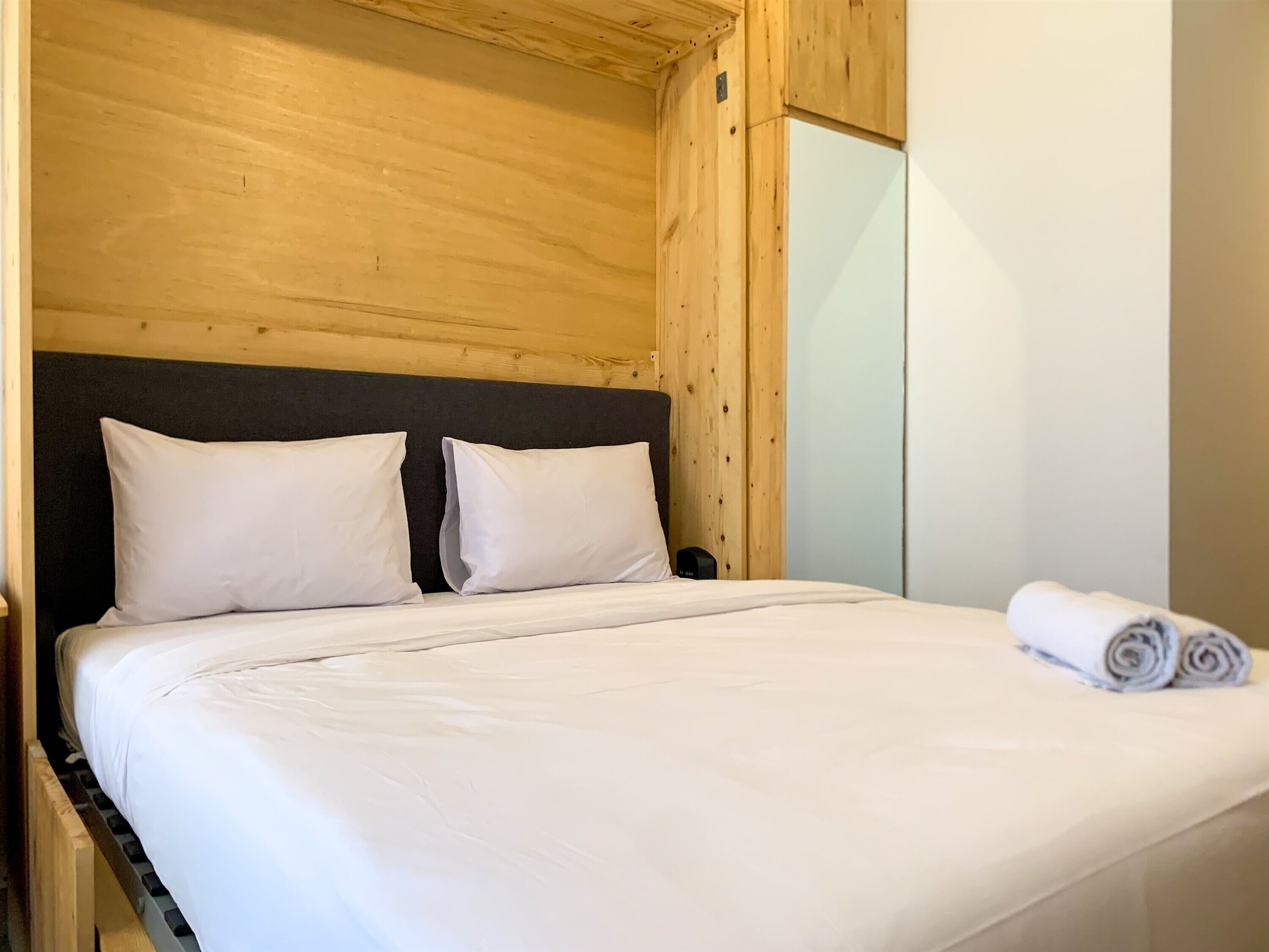 Een bed met witte lakens en twee kussens, een houten hoofdbord, een nachtkastje en een opgerolde handdoek op het bed.