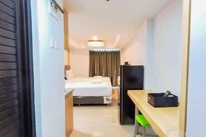 Habitación de hotel con cama, escritorio, silla, refrigerador y ventana con cortinas.
