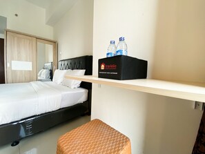Une chambre d’hôtel avec un lit, un petit tabouret, une étagère avec des bouteilles d’eau et une boîte étiquetée « Travelio ».
