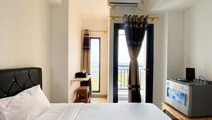 Habitación de hotel con cama, televisor sobre un soporte de madera, equipo de aire acondicionado, ventana con cortinas y vista panorámica de la ciudad.