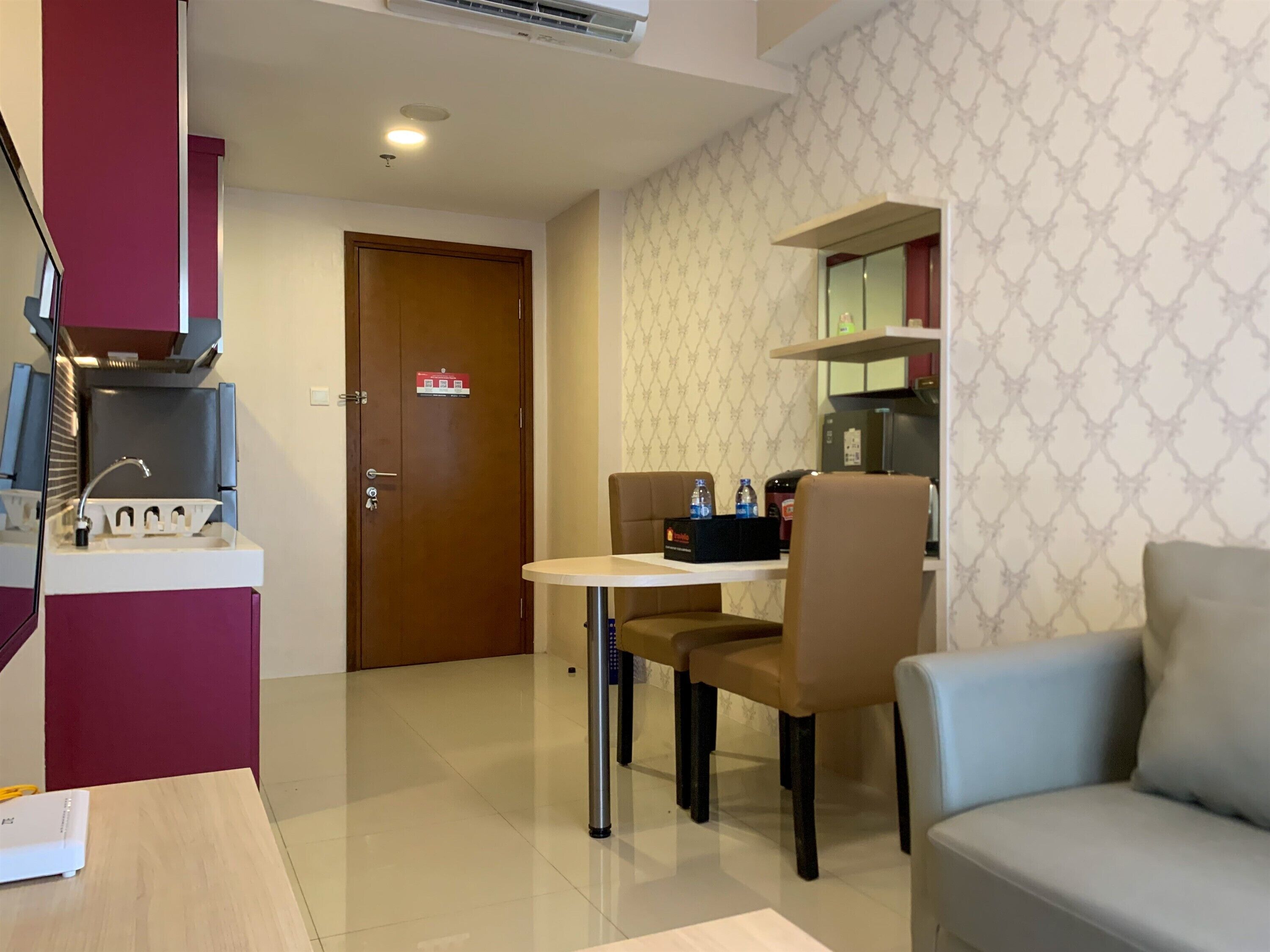 A modern hotel room with a dining area featuring a round table and two chairs, a sofa, a kitchenette, and a door with a fire safety sign.