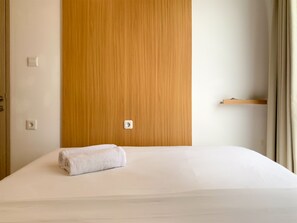 A neatly made bed with a rolled white towel, a wooden wardrobe, and a small shelf against a plain wall. - Strategic And Comfortable 2Br Apartment At Tokyo Riverside Pik 2 (Teluknaga)