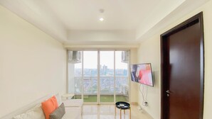 A modern living room with a sofa, a TV, and a view of a cityscape through large windows. - Spacious And Comfort Living 2Br At Menteng Park Apartment (Jakarta)