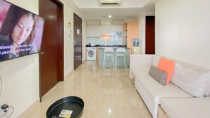 A living room with a white sofa, a flat-screen TV mounted on the wall, and a kitchen area in the background. - Spacious And Comfort Living 2Br At Menteng Park Apartment (Jakarta)