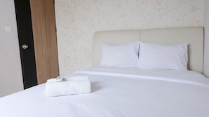 A neatly made bed with white linens and pillows, a folded white towel, and a digital clock on the bed.