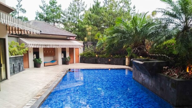 A swimming pool with tiled flooring, surrounded by a landscaped garden with various plants and a building with a tiled roof. - Homey 2Br At Majesty Apartment (Bandung)