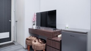 A modern room with a dark wooden desk, a flat-screen TV, a small refrigerator, and two tufted ottomans.