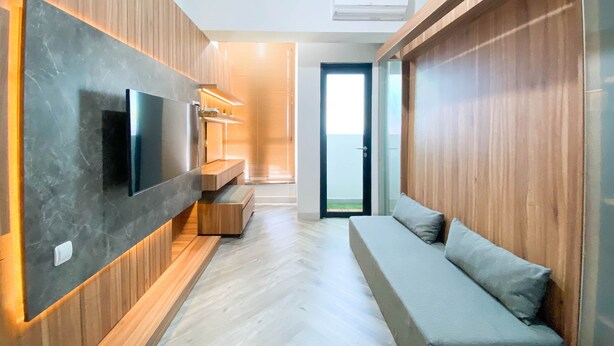 Una sala de estar moderna con televisor empotrado, paneles de madera, un sofá gris y un ventanal corredizo que da a un balcón.