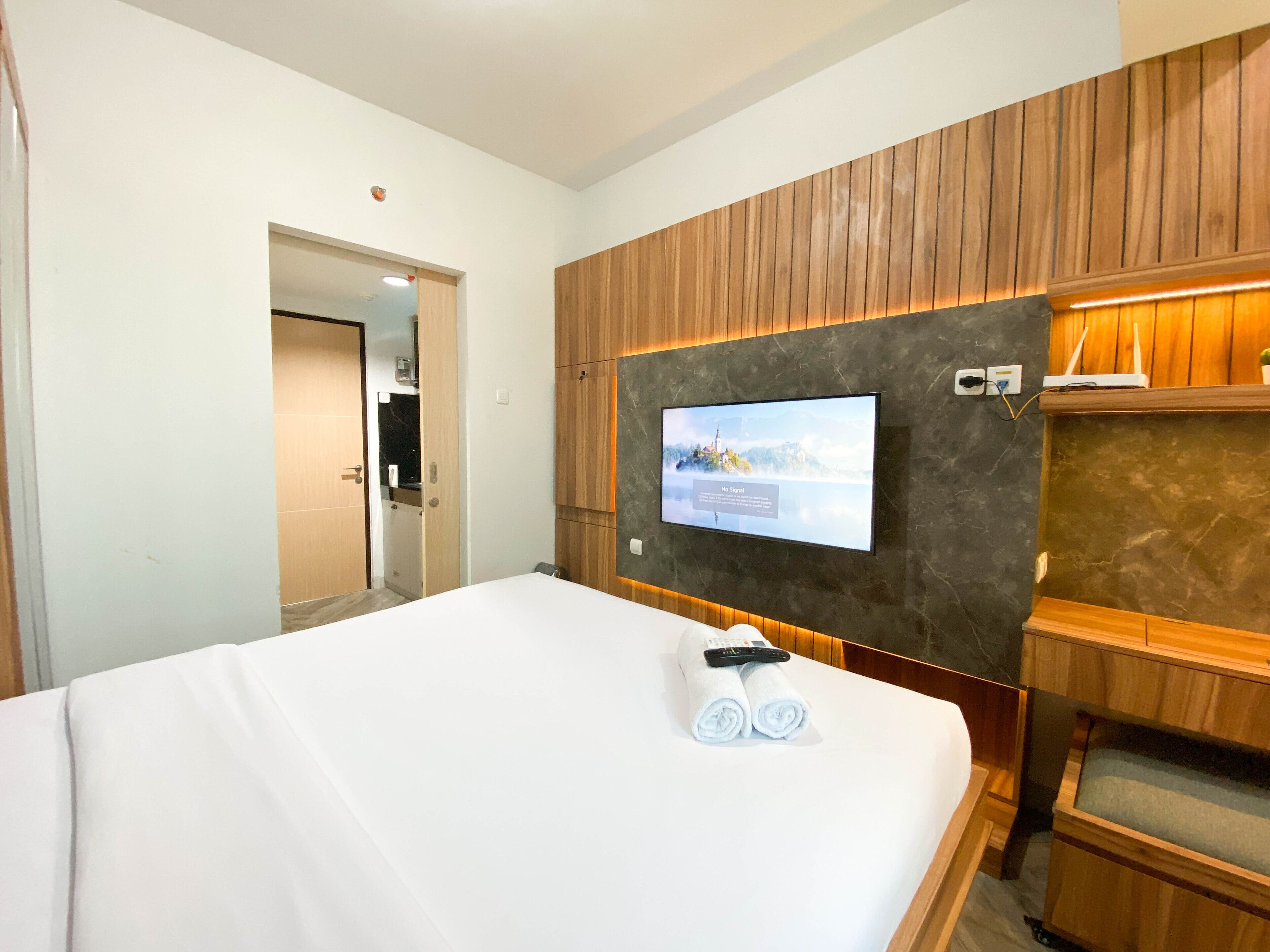 A hotel room with a bed, a television mounted on the wall, wooden paneling, and a small shelf with a remote control and towels.