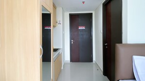 A hotel corridor with a wooden wardrobe, a bed with a white pillow, and a door with a red emergency exit sign. - Comfy And Tidy Studio At Pollux Chadstone Apartment (Cikarang)
