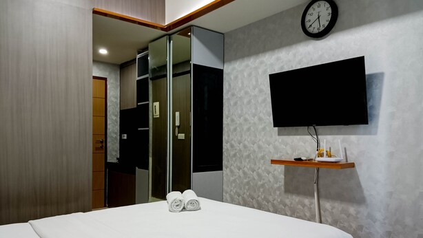 A modern hotel room with a bed, a flat-screen TV mounted on the wall, a clock, and a small shelf with a router and other items. - Relaxing Studio At Vida View Makassar Apartment (Makassar)