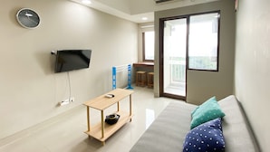 A modern living room with a flat-screen TV, a small wooden table, a sofa with patterned cushions, and a balcony with a view.