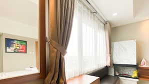 A modern hotel room with a wooden desk, a cushioned bench, a large window with sheer curtains, a marble fireplace, and a wall-mounted TV.