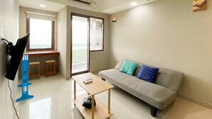 A modern living room with a grey sofa, a small wooden table, and a blue chair. There is a window with a view of greenery outside.