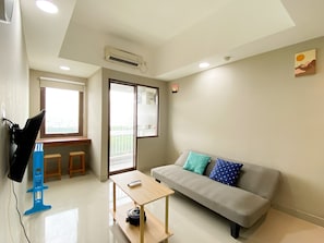 Uma sala de estar moderna com um sofá cinzento, uma pequena mesa de madeira e uma cadeira azul. Há uma janela com vista para vegetação.