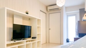 A modern living room with a flat-screen TV on a white entertainment unit, a door, and a window with curtains. - Nice Designed And Elegant 2Br At Green Bay Pluit Apartment (Jakarta)