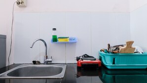 Una cocina con un lavamanos de acero inoxidable, un grifo, un estante con productos de limpieza y un recipiente azul verdoso con utensilios.