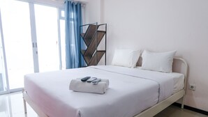 Una cama bien tendida con sábanas blancas y una toalla doblada, una estantería de metal y una ventana con cortinas azules.