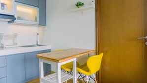 A compact kitchen with a wooden table and yellow chairs, a white shelf with a plant, and a wooden door. - Cozy Stay 1Br At M-Town Signature Apartment (Gading Serpong)