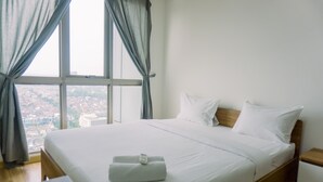 A hotel room with a large bed, a wooden headboard, a bedside table, and a view of a cityscape through a large window with curtains. - Cozy Stay 1Br At M-Town Signature Apartment (Gading Serpong)