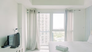 A hotel room with a bed, a TV on a stand, a window with curtains, and a view of buildings outside. - Good Deal And Warm Studio Room Casa De Parco Apartment (South Tangerang)