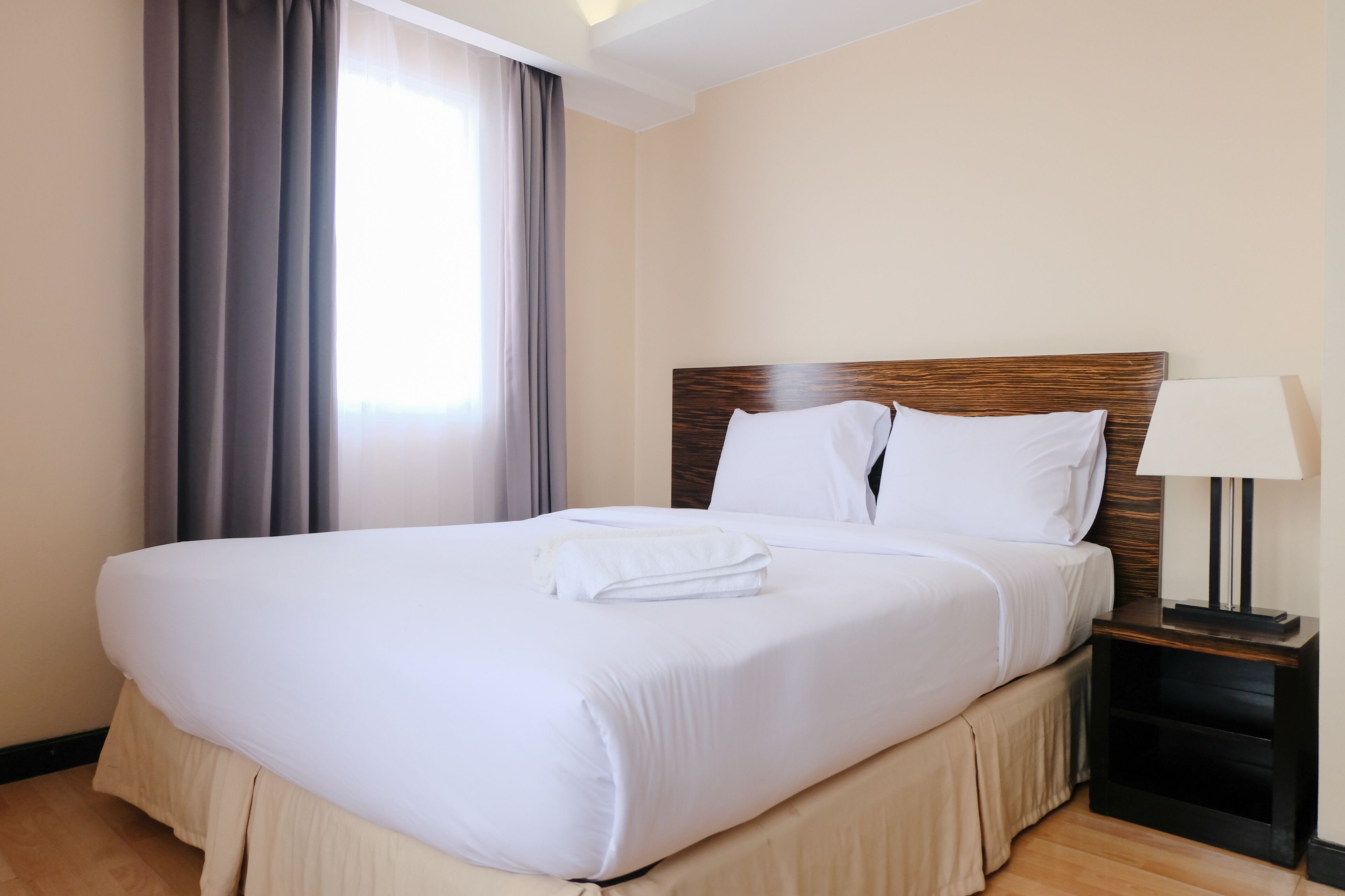 Una cama bien tendida con ropa blanca y una toalla doblada, cabecera de madera, mesita de noche con lámpara y ventana con cortinas.