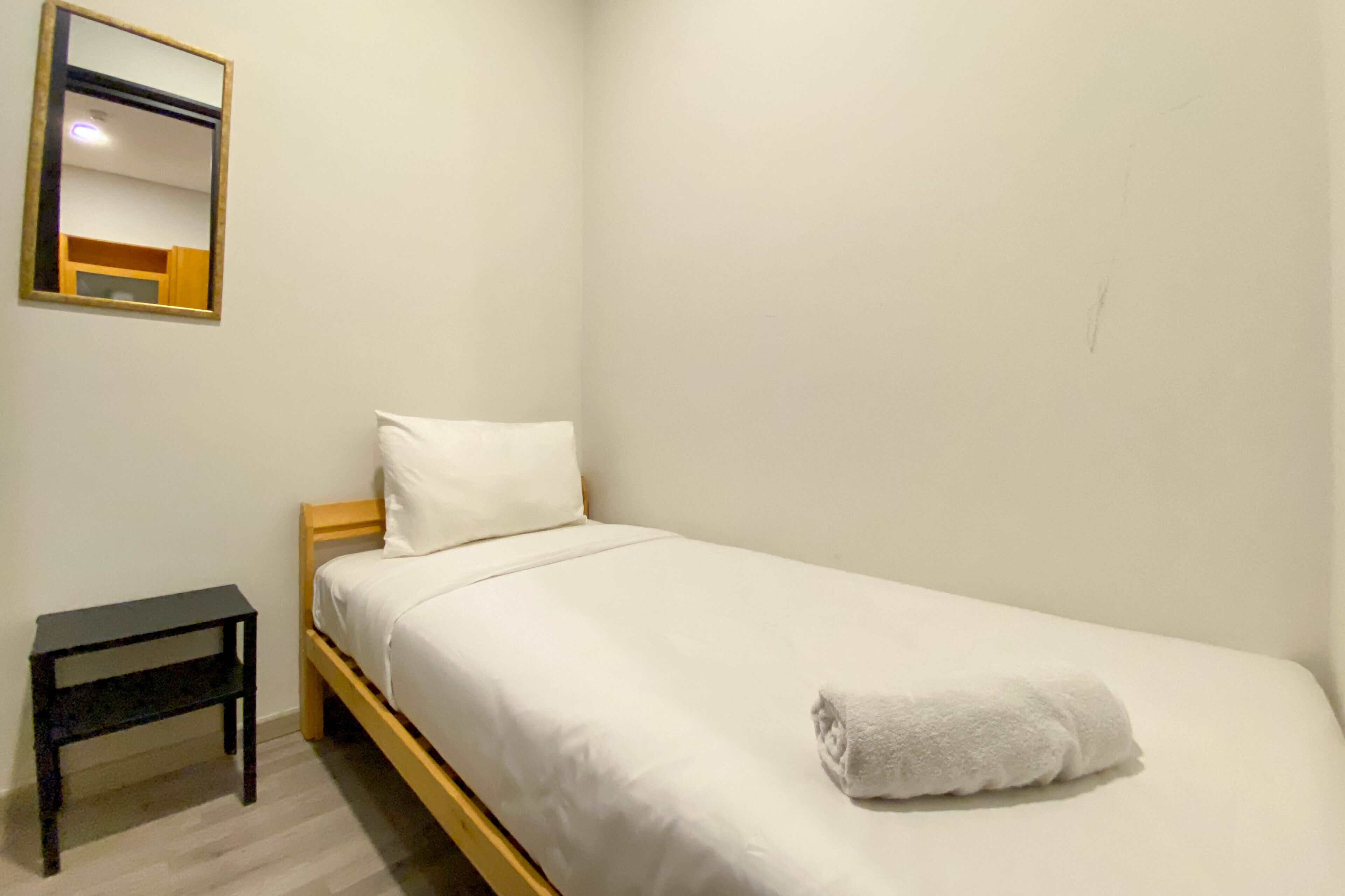 Ein Einzelbett mit weißer Bettwäsche und einem zusammengerollten Handtuch, ein kleiner Nachttisch aus Holz und ein Spiegel an der Wand.