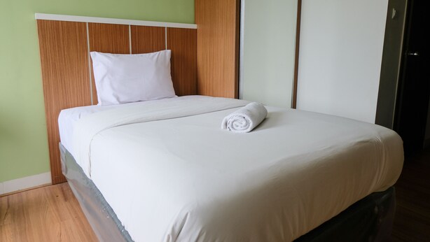 A neatly made bed with a white duvet and a rolled white towel, a wooden headboard, and a green wall. - Best Deal Studio at Harvard Jatinangor Apartment (Cileunyi)