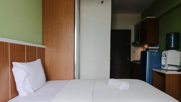 A hotel room with a bed, a wooden headboard, a white pillow, a neatly made bed, a water dispenser, a coffee machine, and a kitchen area with cabinets and a refrigerator. - Best Deal Studio at Harvard Jatinangor Apartment (Cileunyi)
