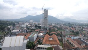 A cityscape with a prominent high-rise building under construction, surrounded by residential buildings and a mountain in the background. - Best Deal Studio at Harvard Jatinangor Apartment (Cileunyi)