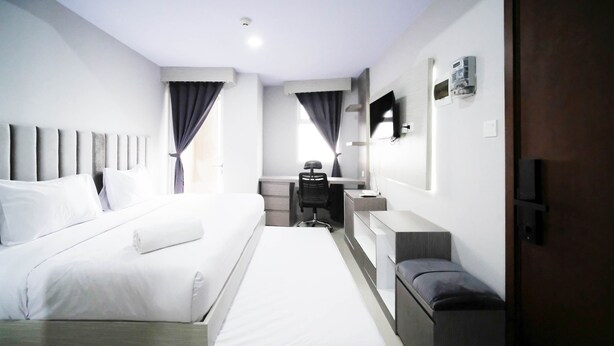 A hotel room with a large bed, a desk, a chair, a TV, and a wardrobe. - Nice And Comfy Studio At Grand Dharmahusada Lagoon Apartment (Surabaya)