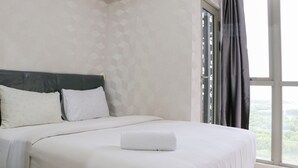 A neatly made bed with white linens and a folded towel, a headboard, and a window with curtains. - Nice And Relaxing 1Br At Gold Coast Apartment (Jakarta)