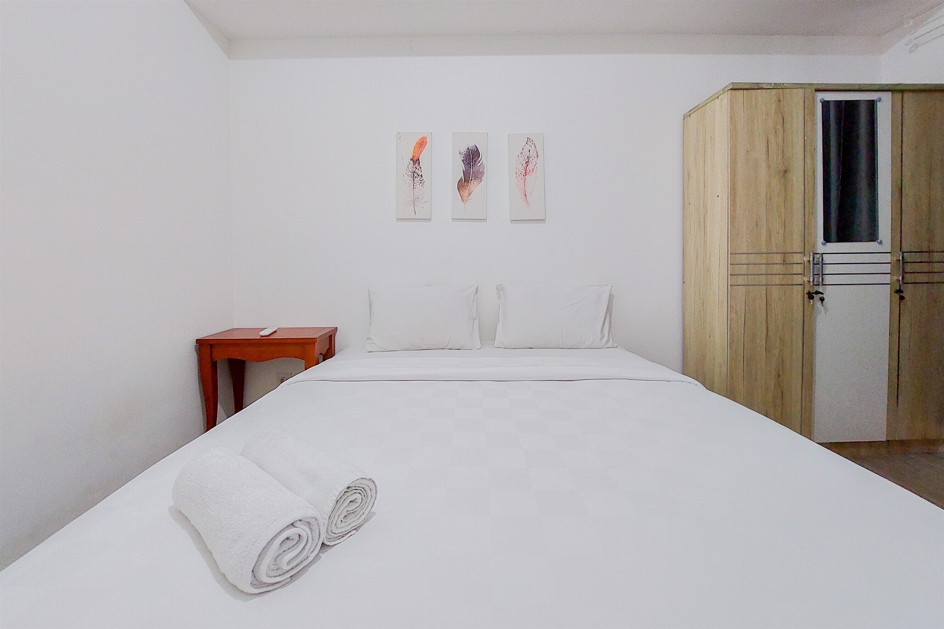 A neatly made bed with white linens and rolled towels, a wooden wardrobe, and three wall-mounted artworks.