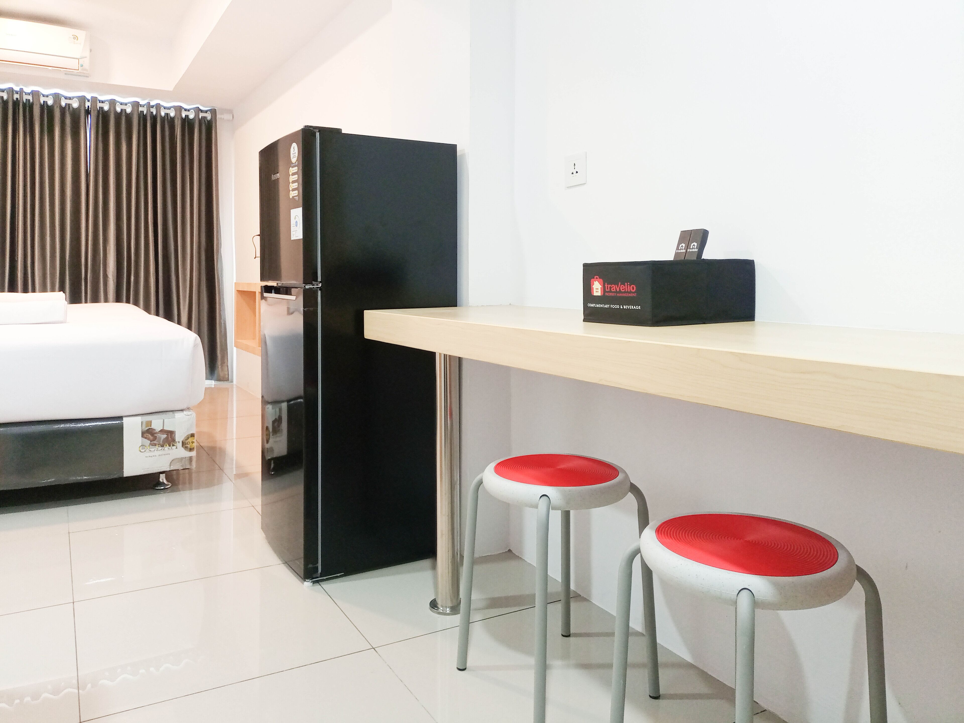 A compact hotel room with a bed, a black refrigerator, a minibar, and two red cushioned stools.