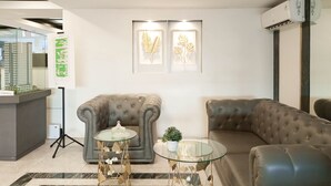 A modern living room with a grey sofa, a glass coffee table, and a decorative plant. There is a framed artwork on the wall and an air conditioning unit mounted on the wall.